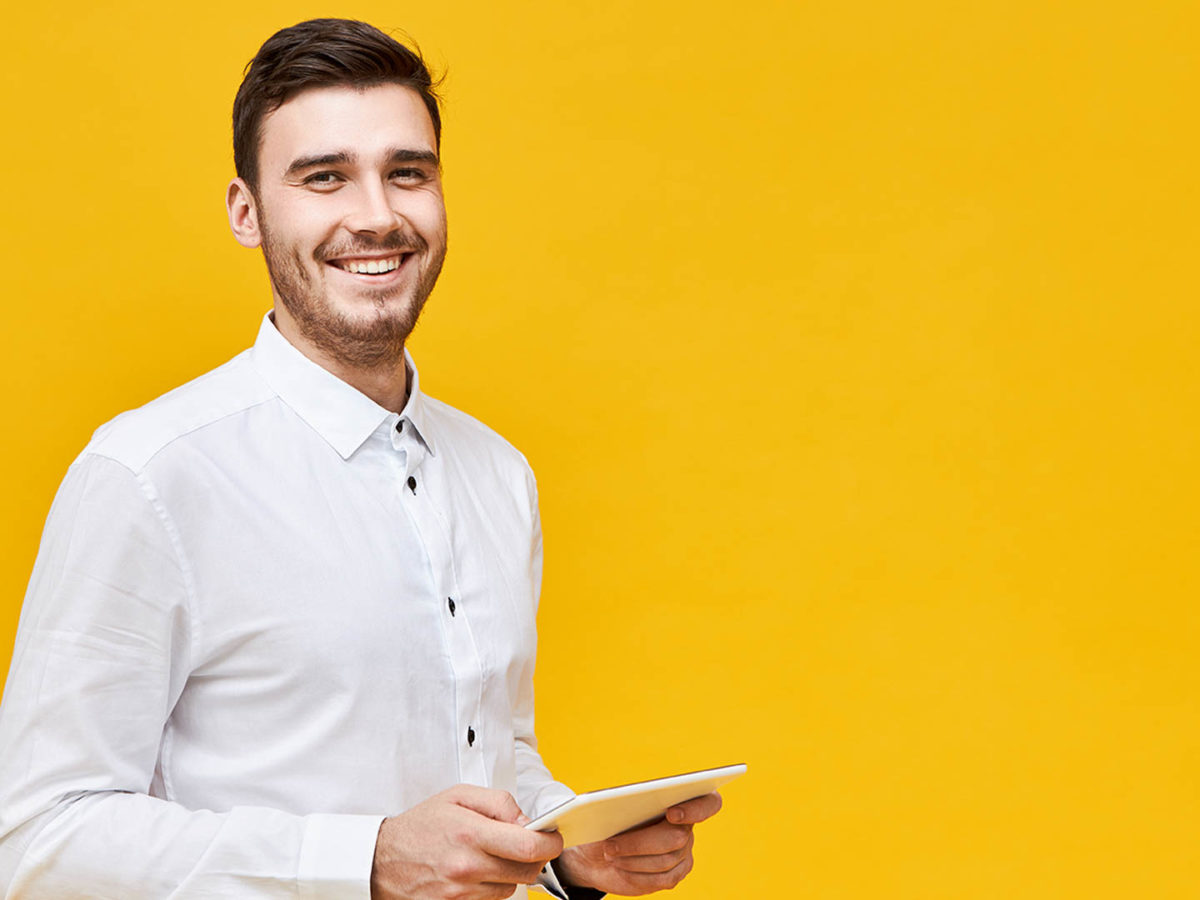 Picture of handsome confident young man in white shirt holding generic digital tablet and smiling broadly, enjoying playing games using online application. Technology, entertainment and gaming