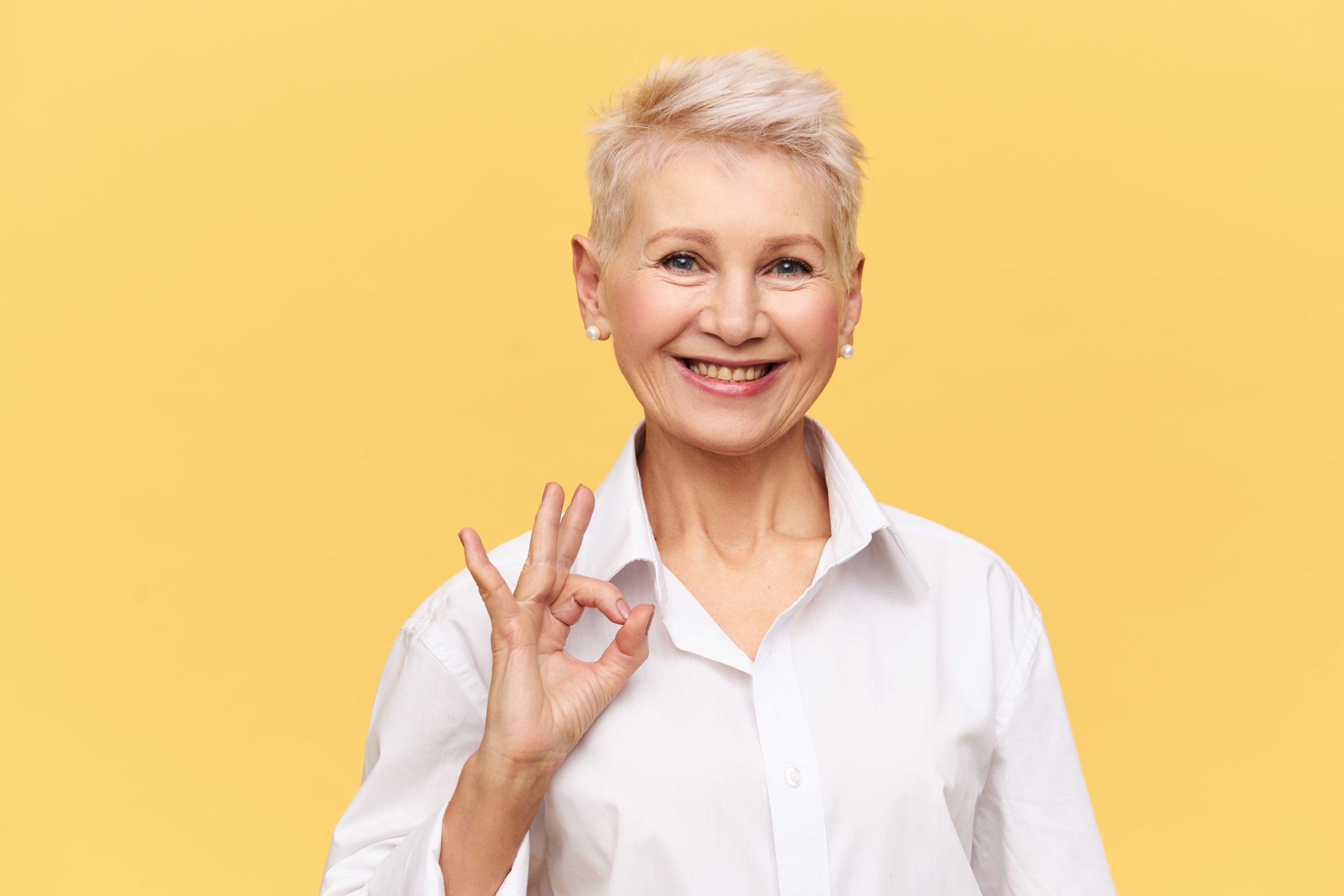 Portrait of successful confident middle aged businesswoman with short dyed hair looking at camera with broad smile making ok gesture, rejoicing at good profitable deal and great yearly income