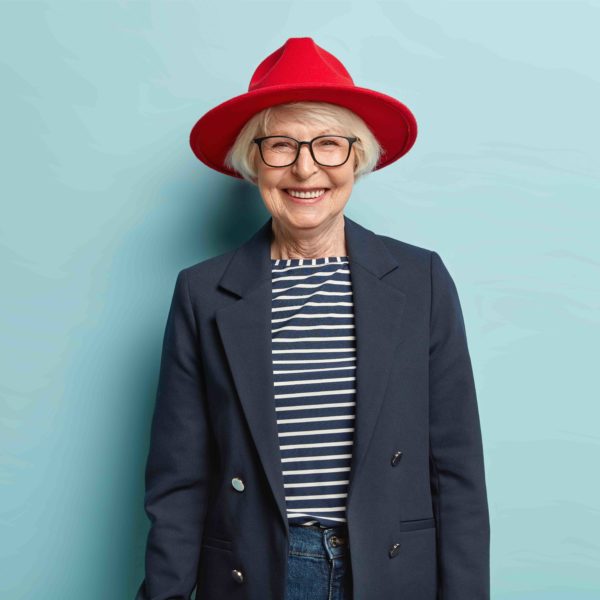 Trendy fashionable elderly woman smiles happily, shows white teeth, has wrinkled skin, dressed in stylish formal clothes, being in good mood, ready for work, enjoys nice day, isolated on blue wall