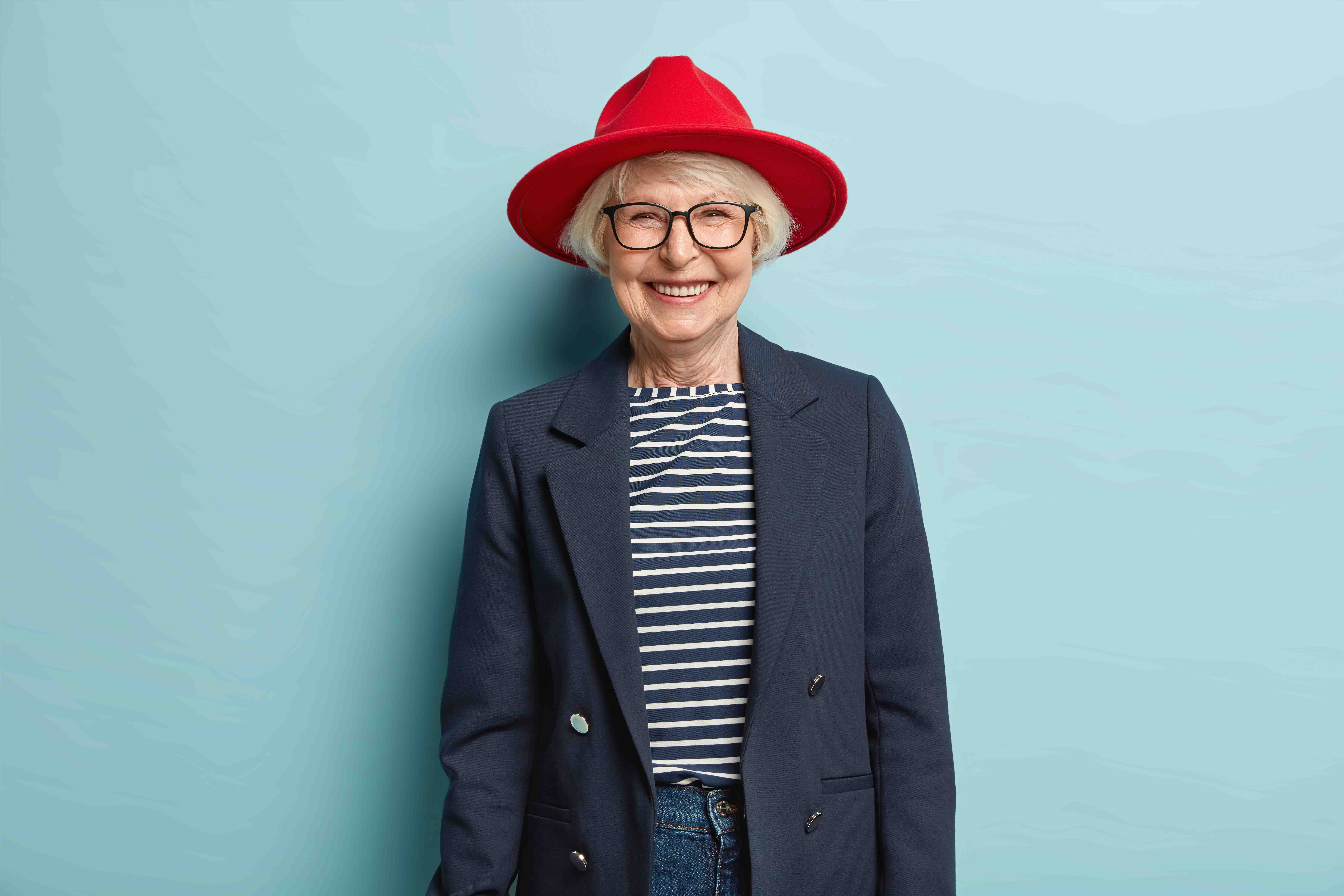 Trendy fashionable elderly woman smiles happily, shows white teeth, has wrinkled skin, dressed in stylish formal clothes, being in good mood, ready for work, enjoys nice day, isolated on blue wall