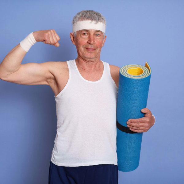 Energetic senior man has physical training, holding yoga mat, showing biceps and his power, looking at camera with pleased expression, warming up body before training.