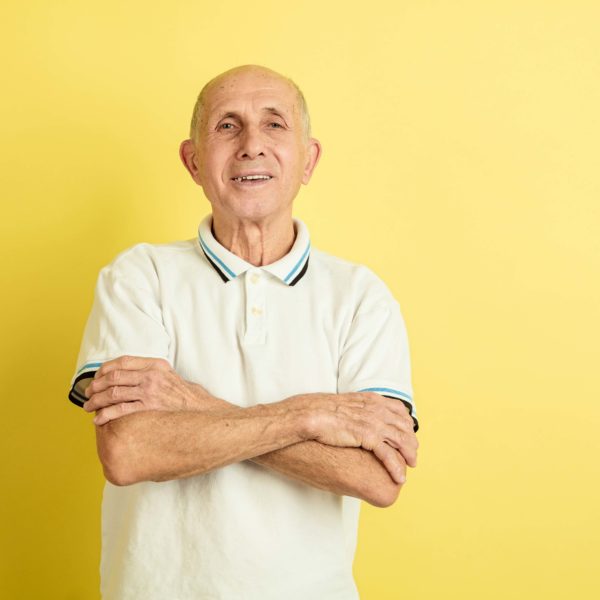 Hands crossed. Caucasian senior man's portrait isolated on yellow studio background. Beautiful male emotional model. Concept of human emotions, facial expression, sales, wellbeing, ad. Copyspace.