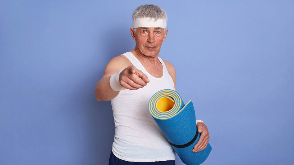 Sporty senior white haired man holding yoga mat, looking directly at camera and pointing with index finger, posing isolated over blue background.,