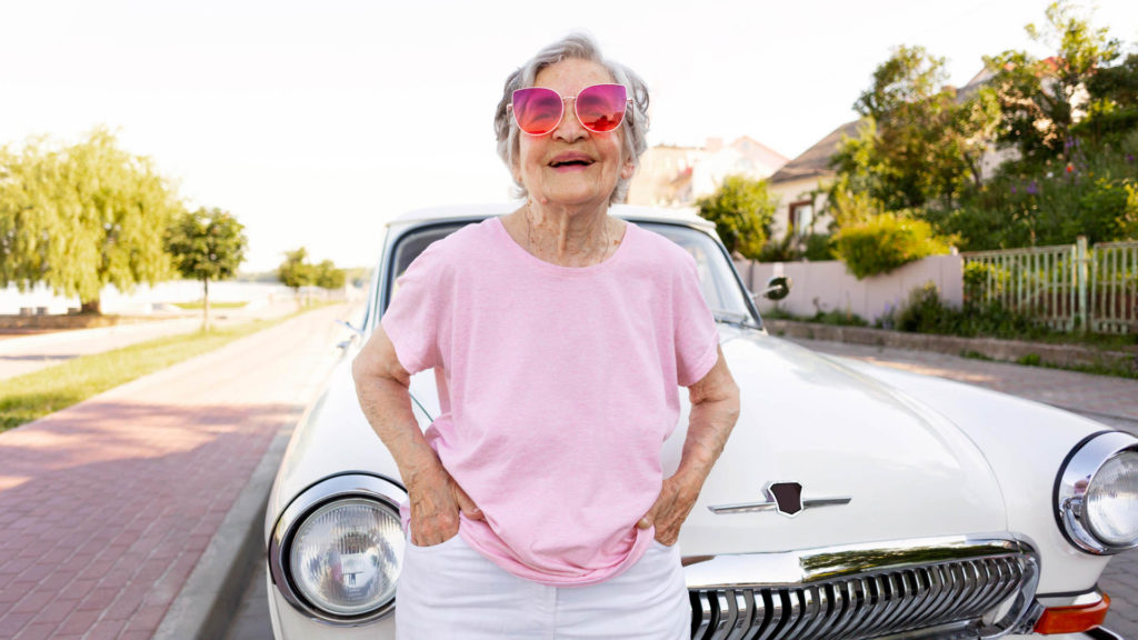older woman wearing pink sunglasses, standing in front of classic car.