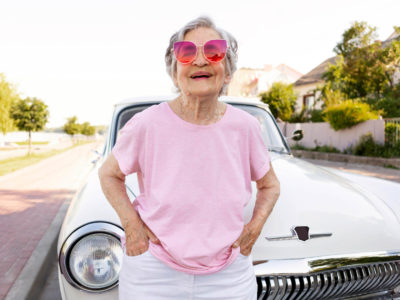 older woman wearing pink sunglasses, standing in front of classic car.
