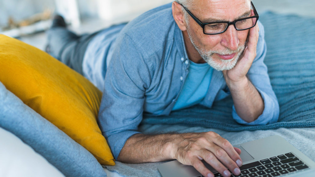 Older man laying on bed looking at laptop and applying for Medicare online.