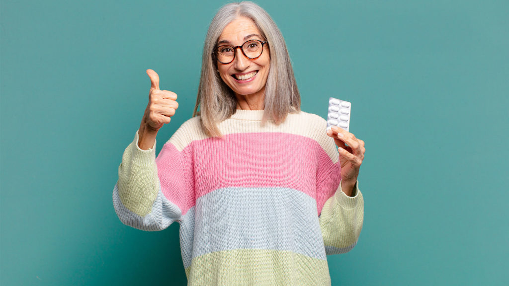 woman with gray hair, smiling with her thumb up, holding a sheet of pills in her other hand that she got from free prescription delivery services.