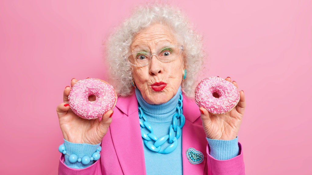 Lovely senior woman holds two glazed donuts keeps lips folded looks pleasantly at camera dressed in fashionable outfit with necklace and bracelet isolated over pink wall. Stylish granny with dessert