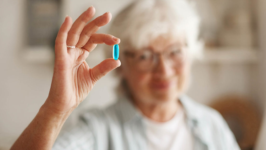 Food, nutrition, diet and health concept. Close up shot of elderly woman's hand holding fish oil or omega-3 polyunsaturated fatty acid supplement in shape of capsule, going to take one during lunch