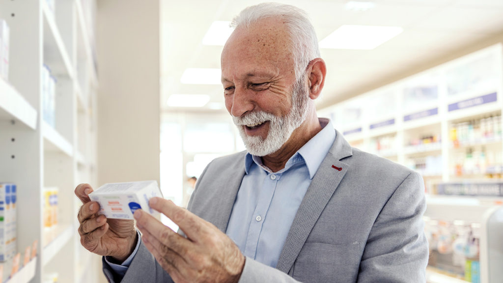 The patient reads the statements on the packaging of the drug at the pharmacy. A senior man with a smile in an elegant suit holds a package of medical drugs and reads the statement and expiration date while considering how to choose a Medicare drug plan.