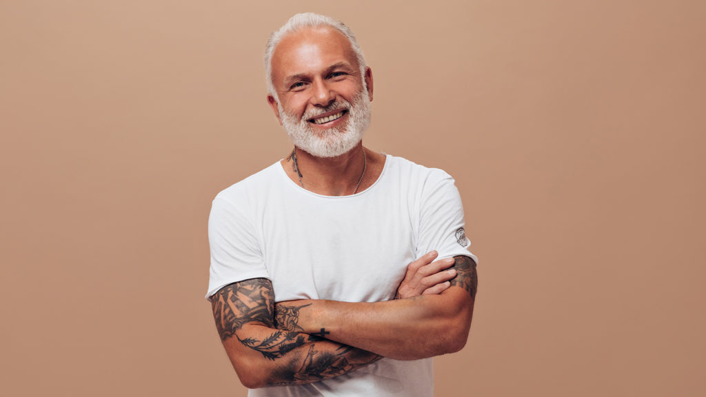Grey haired man in white T-shirt poses on beige background. Handsome guy with beard and tattoos in bright clothes looking into the camera wondering what Medicare does not cover.