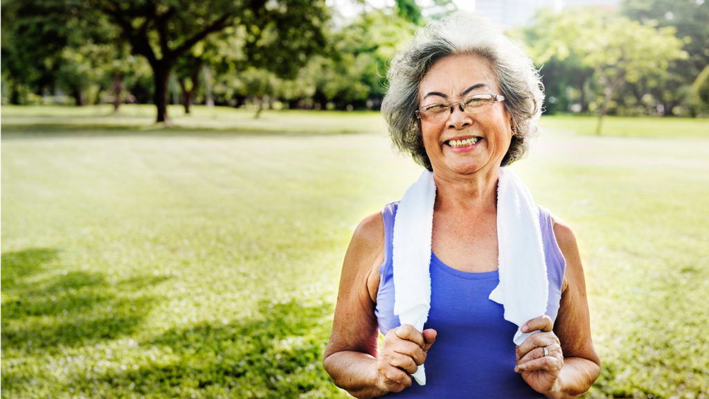 An older Asian American woman smiling in a park-like setting with a workout towel around her neck asking herself, What Does Medicare Cover?