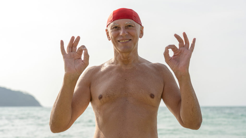 Happy elderly man in a red swimming hat on the beach near the sea water smiling about choosing a Medicare plan.